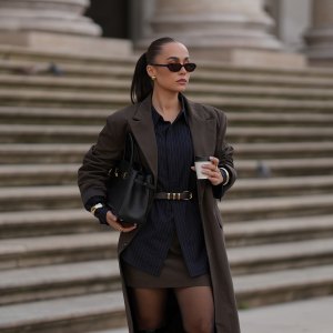 MUNICH, GERMANY - SEPTEMBER 16: Anna Wein is seen wearing a long brown coat with long sleeves and pinstripe details from Lounge; a dark pinstriped shirt with black buttons; a short brown skirt with subtle pinstripes from Lounge; sheer black tights; tall black leather heeled boots with a chunky heel and a square toe from H&M; a structured black leather handbag with gold hardware; a black leather belt, tied around the waist, with a golden fastener; slim dark sunglasses with brown tinted lenses from Corlin Eyewear; subtle golden metallic jewelry featuring rings and bracelets from Mango; her brown hair is styled in a sleek ponytail on September 16, 2025 in Munich, Germany. (Photo by Moritz Scholz/Getty Images)