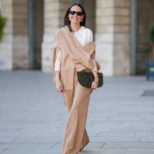 Alba Garavito Torre wears black sunglasses from Ray Ban, white large pearls earrings, a white shirt with cut-out lace pattern large collar, a beige felt / wool oversized cloak, matching beige wool / felt palazzo pants, a black shiny leather ruffled handbag, a gold Love bracelet from Cartier, gold bracelet, gold rings, brown leather pointed pumps heels shoes, during a street style fashion photo session, on October 28, 2021 in Paris, France.
