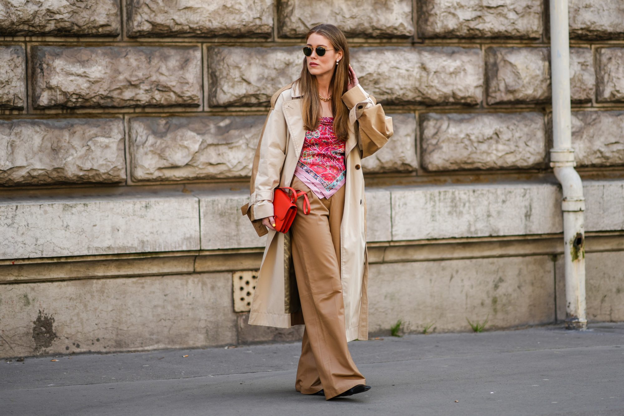 PARIS, FRANCE - MARCH 05: Annabel Rosendahl wears sunglasses, pearls pendant earrings, a gold large chain necklace, a beige and brown long oversized coat, a red shiny leather Constance shoulder bag from Hermes, a red / pink / green / black print pattern silk scarf as a shoulder-off t-shirt, brown shiny leather wide legs pants, black shiny leather pointed pumps heels shoes, outside Hermes , during Paris Fashion Week - Womenswear F/W 2022-2023, on March 05, 2022 in Paris, France. (Photo by Edward Berthelot/Getty Images)