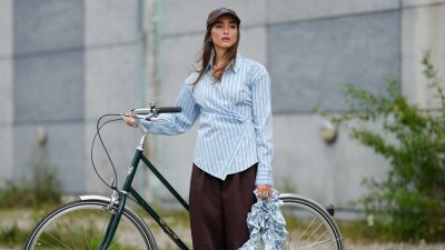 COPENHAGEN, DENMARK - AUGUST 07: A guest wears a brown baseball cap with embroidered details. Hair is styled straight and loose. The outfit features a light blue button-up shirt with vertical dark blue stripes, designed with an asymmetrical wrap detail. Paired with wide-legged, dark brown trousers. Footwear consists of blue sandals. Accessories include a ruffled, striped bag in shades of blue and white, outside Marimekko, during Copenhagen Fashion Week, on August 07, 2025 in Copenhagen, Denmark (Photo by Edward Berthelot/Getty Images)
