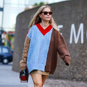 A guest wears sunglasses, earrings, a brown and blue knitted wool pullover/dress with red v-neck, a bag, during London Fashion Week September 2019 on September 14, 2019 in London, England.