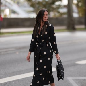 A Fashion Week guest is seen wearing round diamond stud earrings; a long-sleeved black midi dress with white polka dot pattern, notched lapels, and a tie belt; a gold bangle; a rose gold and black watch from Rolex; a gold ring; and a rectangular black handbag with a white polka dot pattern before the Elie Saab Womenswear Spring/Summer 2025 Show as part of Paris Fashion Week on September 28, 2024 in Paris, France.
