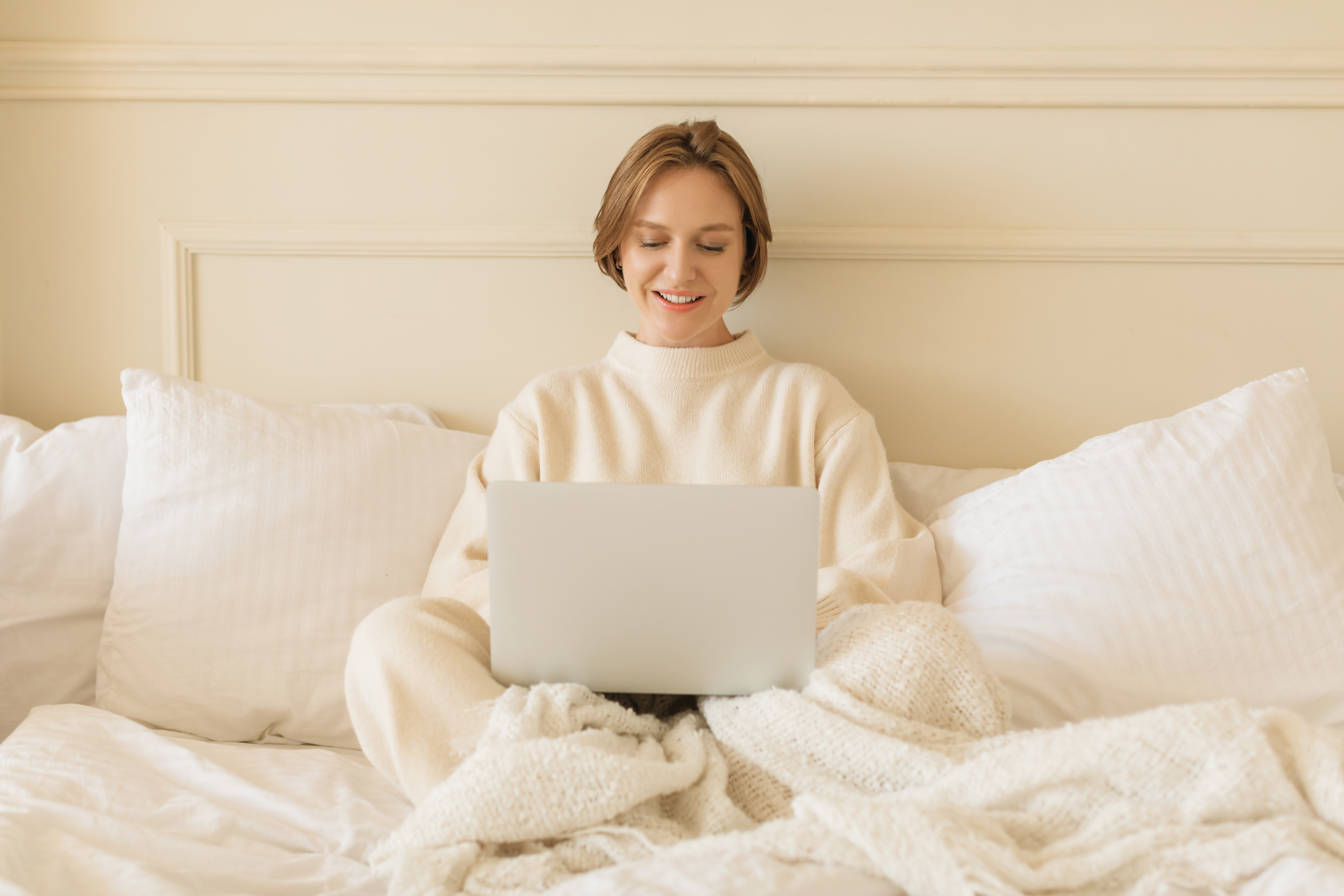 Smiling young woman in casual loungewear sitting on her bed working on her laptop computer