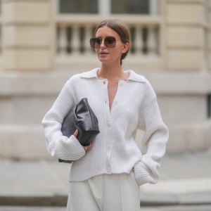 Diane Batoukina wears a white ribbed knit cardigan with a wide collar and oversized sleeves by Lovechild 1979, paired with high-waisted ivory trousers by Herskind. A dark brown leather clutch bag by St. Agni is held under one arm. Oversized square Dior sunglasses with brown gradient lenses are worn. Hair is parted at the center and styled in a sleek low bun. Natural makeup with even skin and soft pink lips is visible. Mid shot, during a street style fashion photo session, on April 17, 2025 in Paris, France.