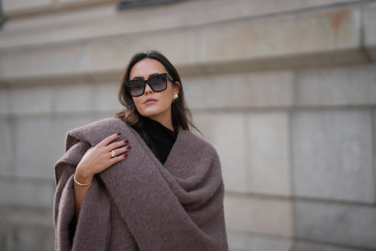 Laura Noltemeyer is seen wearing an oversized fringed wool poncho in a mauve tone with a textured, fluffy finish, fringes at the hem and a gold pin closure element called „Poncho COZY“ by Aylin Koenig; black fine-knit turtleneck sweater with slim-fitting silhouette by Aylin Koenig; among her jewelry is a white gold „LOVE“ bracelet by Cartier; oversized square black acetate sunglasses with ombré tinted lenses by Céline; large gold statement earrings with a ruffled surface and a green stone element by Mango; her dark brown hair is styled in loose waves with a center part on November 13, 2025 in Hamburg, Germany.