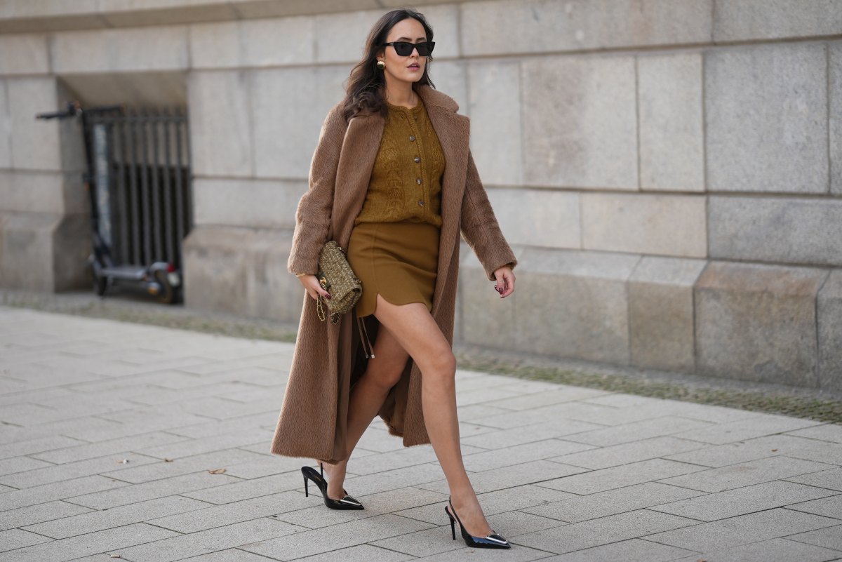 Laura Noltemeyer is seen wearing a a long camel-colored brushed wool coat with a wrap silhouette and integrated belt by Max Mara; a mustard-colored knit cardigan with a brushed cable-knit pattern and front button fastening by Sézane; a mustard yellow brownish mini skirt in a soft wool fabric by Sézane; black patent leather slingback pumps with a sharply pointed toe and silver hardware accents by Yves Saint Laurent; a small quilted tweed flap bag in a khaki Color with woven fabric material with a gold chain strap and CC clasp by Chanel; gold statement earrings with a ruffled surface and a green stone element by Mango; black cat eye sunglasses with an acetate frame by Yves Saint Laurent; her brown hair is styled in loose waves with a center part on November 13, 2025 in Hamburg, Germany.