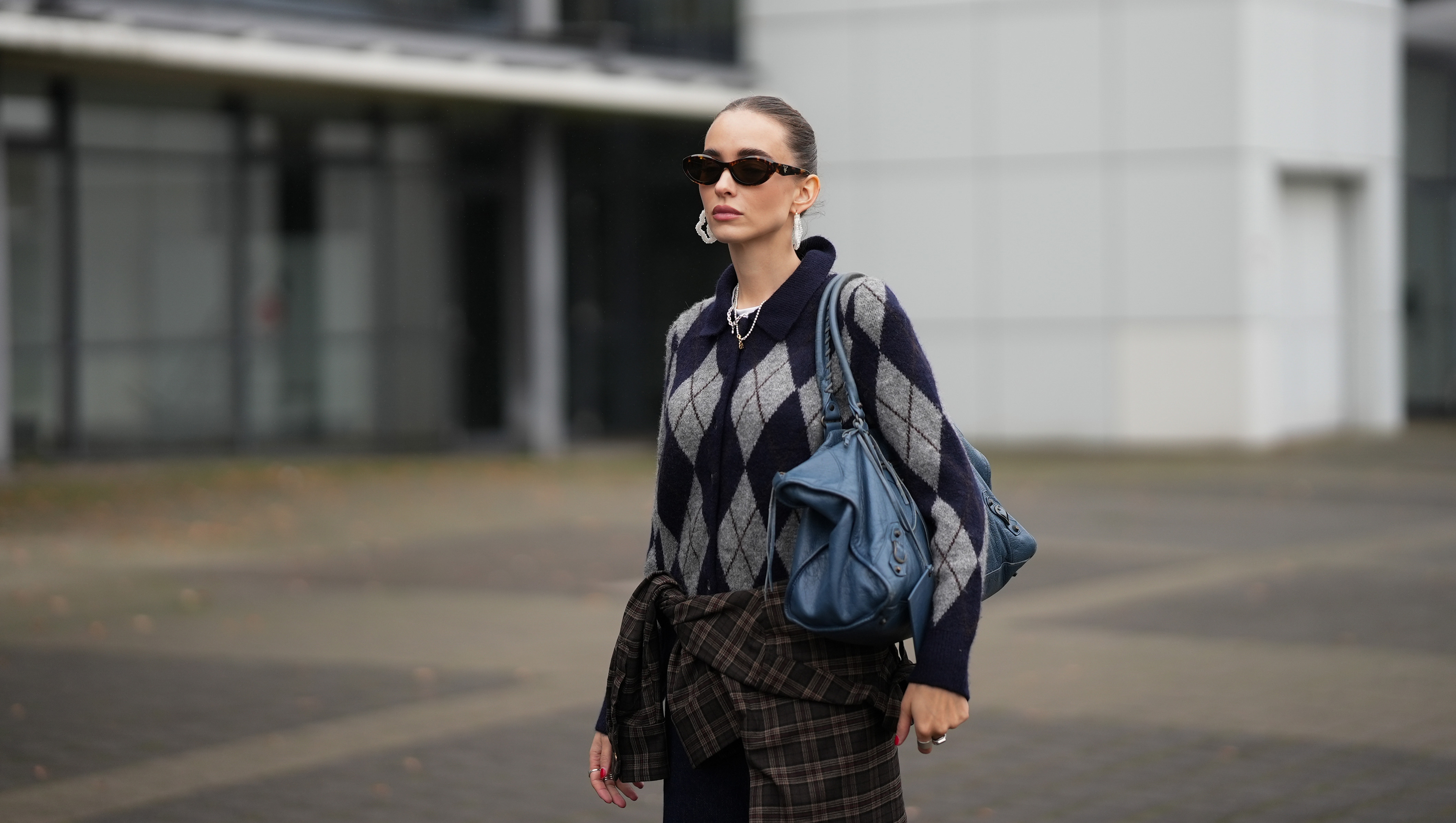 DUESSELDORF, GERMANY - OCTOBER 14: Evi Wave is seen wearing a navy blue and grey argyle knit H&M cardigan with a collared neckline, front button closure with tonal buttons and ribbed cuffs; some delicate chain and ball chain necklaces; a plaid Our Legacy overshirt in brown-based tartan pattern is tied around the waist; on her fingers she wears silver rings from Ariane Ernst and her nails are polished in red; she carries a navy blue Balenciaga Le City bag maxi in Arena Storico lambskin with two leather hand-braided handles, brass hardware, front zipped pocket and knotted leather puller; her brown hair is slicked back into a neat low bun; she wears a pair of cat-eye Prada sunglasses in tortoiseshell acetate with dark lenses; she wears a pair of sculptural beaded earrings handmade with white glass beads by Kroon02 on October 14, 2025 in Duesseldorf, Germany. (Photo by Moritz Scholz/Getty Images)
