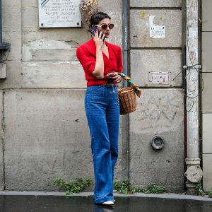 A guest wears circle sunglasses, a red fluffy puffy short sleeves / buttoned cropped cardigan, navy blue denim wide legs pants, a brown wickers basket handbag, a green / blue / orange print pattern silk scarf, rings, white shiny varnished leather heels shoes, a gold chain belt, outside the Hermes show, during Paris Fashion Week - Menswear Spring/Summer 2023, on June 25, 2022 in Paris, France.
