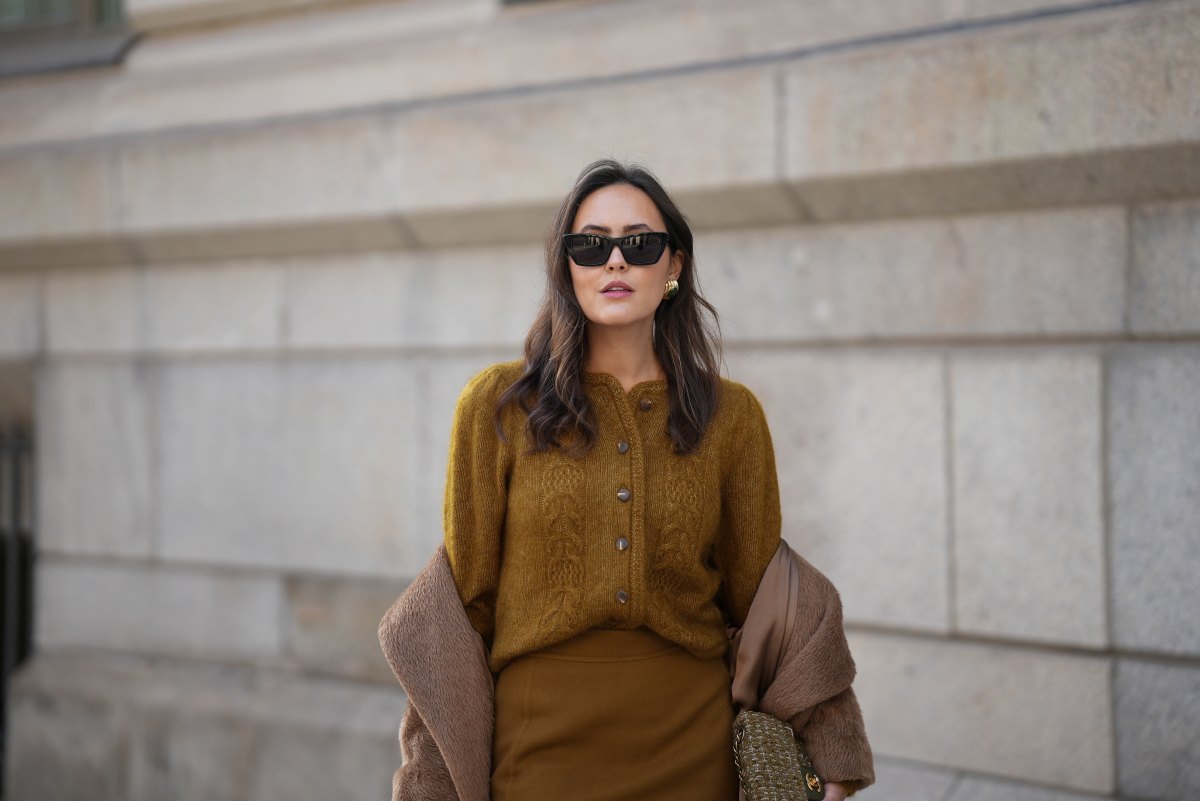 Laura Noltemeyer is seen wearing a a long camel-colored brushed wool coat with a wrap silhouette and integrated belt by Max Mara; a mustard-colored knit cardigan with a brushed cable-knit pattern and front button fastening by Sézane; a small quilted tweed flap bag in a khaki Color with woven fabric material with a gold chain strap and CC clasp by Chanel; gold diamond rings by Ariane Ernst; gold statement earrings with a ruffled surface and a green stone element by Mango; black cat eye sunglasses with an acetate frame by Yves Saint Laurent; her brown hair is styled in loose waves with a center part on November 13, 2025 in Hamburg, Germany.