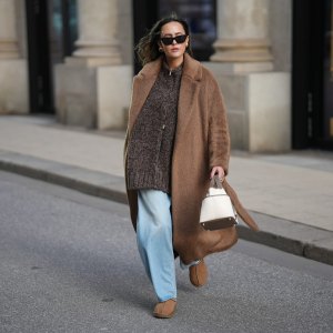 HAMBURG, GERMANY - NOVEMBER 13: Laura Noltemeyer is seen wearing a long camel-colored brushed wool wrap coat with a belt and wide lapels by Max Mara; a chunky zip-front sweater cardigan in a mélange brown wool blend with a textured, hand-knit look by by Aylin Koenig; light blue wide-leg jeans in a soft washed denim with a relaxed fit and front tag detail by Closed; chestnut colored suede slippers with plush lining called „Tasman“ by UGG; a structured bucket-shaped handbag in an off-white linen shade with taupe leather handles and lining details called „Picotin Cargo 18 Toile Goeland“ by Hermès; black cat eye sunglasses with an acetate frame by Yves Saint Laurent; gold statement earrings with a ruffled surface and a green stone element by Mango ; her brown hair is worn loose in soft waves with a center part on November 13, 2025 in Hamburg, Germany. (Photo by Moritz Scholz/Getty Images)