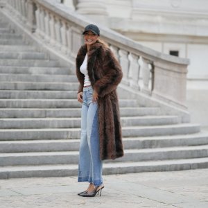 PARIS, FRANCE - DECEMBER 11: Alicia Aylies wears hair pulled back into a sleek low ponytail, natural skin with defined brows and lipstick, a black baseball NY cap, a white fitted cropped top by Celine, a long brown faux fur coat with a straight cut and button closure, light blue high waist flared wide leg jeans with a raw hem, and dark pointed toe high heel pumps shoes, during a street style fashion photo session, on December 11, 2025 in Paris, France. (Photo by Edward Berthelot/Getty Images)