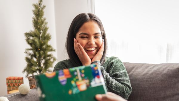 Young woman sitting on the sofa. Her boyfriend is giving her a present for Christmas