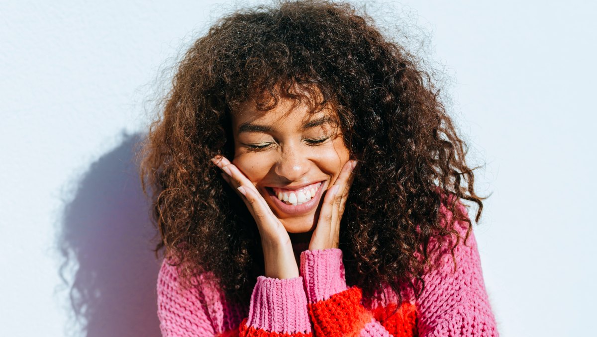 Portrait of a cheerful young woman with curly hair wearing colorful sweater, touching her face, smiling and closing her eyes