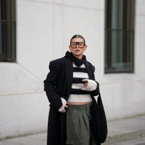 BERLIN, GERMANY - NOVEMBER 25: Sophia Geiss is seen wearing an oversized long black wool coat with structured shoulders and a straight, ankle-length silhouette from Herskind; a slightly cropped, fuzzy black-and-white striped knit sweater from H&M; wide-leg khaki green cargo trousers with drawstring waist and large side pockets from The Frankie Shop; a black suede crescent shoulder hobo bag with a minimalist curved shape from Aesther Ekme; oversized aviator glasses with tortoiseshell frame from Prada; a black knit balaclava from COS; small silver hoop earrings from Maria Black; her brown hair is styled in a sleek low bun with a center parting on November 25, 2025 in Berlin, Germany. (Photo by Moritz Scholz/Getty Images)