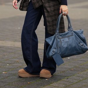DUESSELDORF, GERMANY - OCTOBER 14: Evi Wave is seen wearing a navy blue and grey argyle knit H&M cardigan with a collared neckline, front button closure with tonal buttons and ribbed cuffs; some delicate chain and ball chain necklaces; a plaid Our Legacy overshirt in brown-based tartan pattern is tied around the waist; a pair of dark indigo wide-leg Closed jeans with a relaxed fit; her shoes are Chunky platform suede UGG mini boots in a camel-tan color; on her fingers she wears silver rings from Ariane Ernst and her nails are polished in red; she carries a navy blue Balenciaga Le City bag maxi in Arena Storico lambskin with two leather hand-braided handles, brass hardware, front zipped pocket and knotted leather puller; her brown hair is slicked back into a neat low bun; she wears a pair of cat-eye Prada sunglasses in tortoiseshell acetate with dark lenses; she wears a pair of sculptural beaded earrings handmade with white glass beads by Kroon02 on October 14, 2025 in Duesseldorf, Germany. (Photo by Moritz Scholz/Getty Images)
