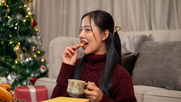A cheerful, beautiful Asian woman in a cozy red sweater enjoys Christmas cookies and a warm drink while reading in her living room, spending the Christmas holiday peacefully at home alone.