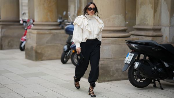 Jennifer Casimiro is seen wearing a cream cable-knit turtleneck sweater with voluminous ruffled detailing on the shoulders from Zara; black high-waisted trousers with a tapered silhouette, cinched at the ankles from Zara; black lace-up shoes with block heels, featuring pom-pom accents at the ankle ties from Gucci; the Suede Varenne small black clutch bag with a rectangular shape and embossed gold branding from Jimmy Choo; oversized black rectangular sunglasses from MiuMiu; gold cuffed earrings from Zara; her black hair is worn down with a center part on September 19, 2025 in Hamburg, Germany.