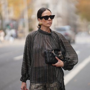 Patricia Wirschke is seen wearing a shimmering metallic striped blouse in shades of charcoal, bronze, and silver with a see through, flowing silhouette, high neckline and tie detail at the collar by Si Yu; paired with satin wide-leg trousers featuring an intricate geometric print in gold and dark brown tones by Si Yu; a small black quilted leather “Lady D-Joy” handbag with gold hardware by Dior; black cat-eye sunglasses with a chunky rim by Phoebe Philo; gold statement hoop earrings by an unknown brand; a rose gold “B.zero1” ring by Bulgari; a white gold “Alhambra” bracelet by Van Cleef & Arpels; her brown hair is in a sleek low bun parted in the middle on November 10, 2025 in Duesseldorf, Germany.
