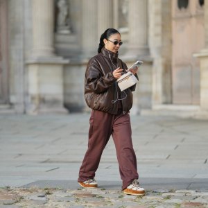 PARIS, FRANCE - DECEMBER 31: Yuwei Zhangzou wears hair pulled back into a low braided ponytail, makeup with an even complexion, defined brows and lipstick, wire earphones, oval dark-lens silver sunglasses by Celine, a brown oversized bomber leather jacket with a high collar and front zip closure, straight-leg brown sport jogger pants by Alo, a small structured beige and brown shearling fluffy bag with a top handle by Loro Piana, white shearling-lined lace-up shoes with brown suede panels, contrast laces and a flat rubber sole by Moon Boot, holds a mobile phone, during a street style fashion photo session, on December 31, 2025 in Paris, France. (Photo by Edward Berthelot/Getty Images)