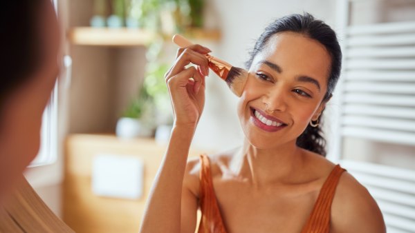 Beautiful girl applying makeup using powder brush before going to work. Healthy latin woman looking in the mirror and applying cosmetic with a big brush. Young woman looking in the mirror and applying foundation or blusher on her face.