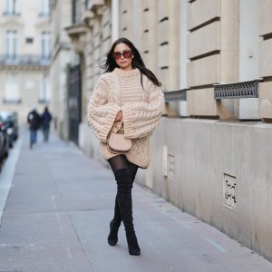 PARIS, FRANCE - DECEMBER 09: Patricia Gloria Contreras wears long straight dark brown hair worn loose, natural skin with neutral lipstick, oversized square burgundy tinted sunglasses, an oversized beige chunky knit sweater dress with a ribbed crew neckline, long ribbed cuffs and a textured cable knit pattern, sheer black tights, black suede over the knee boots, and a beige leather small top handle bag with a gold metal handle by Chloe, during a street style fashion photo session, on December 09, 2025 in Paris, France. (Photo by Edward Berthelot/Getty Images)