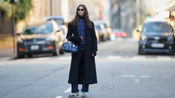 PARIS, FRANCE - DECEMBER 27: Amanda Derhy wears long curly / wavy brown hair worn loose with a center part, black oval sunglasses from Miu Miu, make-up is a natural complexion with defined brows and soft pink lipstick, a black wool winter long coat, a black ribbed turtleneck top layered under a dark indigo blue denim jacket with a pointed collar, vertical seam detailing and a fitted silhouette, a dark blue leather Coach bag with metal ring handles and visible logo plaque, matching straight-leg dark indigo denim jeans pants, Adidas SL72 OG sneakers shoes in white with green shoelaces and contrasting black detailing / cow pattern print, during a street style fashion photo session, on December 27, 2025 in Paris, France. (Photo by Edward Berthelot/Getty Images)