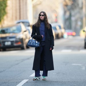 PARIS, FRANCE - DECEMBER 27: Amanda Derhy wears long curly / wavy brown hair worn loose with a center part, black oval sunglasses from Miu Miu, make-up is a natural complexion with defined brows and soft pink lipstick, a black wool winter long coat, a black ribbed turtleneck top layered under a dark indigo blue denim jacket with a pointed collar, vertical seam detailing and a fitted silhouette, a dark blue leather Coach bag with metal ring handles and visible logo plaque, matching straight-leg dark indigo denim jeans pants, Adidas SL72 OG sneakers shoes in white with green shoelaces and contrasting black detailing / cow pattern print, during a street style fashion photo session, on December 27, 2025 in Paris, France. (Photo by Edward Berthelot/Getty Images)