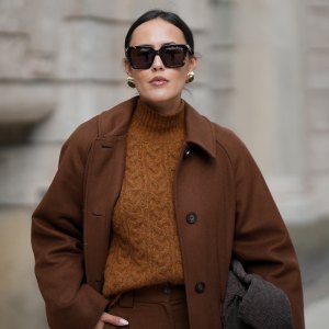 HAMBURG, GERMANY - OCTOBER 16: Laura Noltemeyer is seen wearing a long straight tailored wool coat in chocolate brown with button fastening by Sézane; high-waisted wide-leg pleated pants by Sézane; a textured cable knit sweater in a brown tone by Sézane; a suede asphalt grey shoulder bag with a woven leather structure by Les Visionnaires; square brown sunglasses with gold detailing by Bottega Veneta; gold statement earrings with a green stone detail by Mango; her dark hair is in a low ponytail on October 16, 2025 in Hamburg, Germany. (Photo by Moritz Scholz/Getty Images)