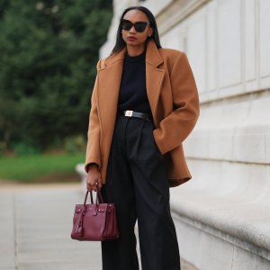 PARIS, FRANCE - DECEMBER 13: Emilie Joseph wears long straight dark brown hair parted at the center, makeup featuring an even complexion with matte skin and deep red lipstick, oversized black sunglasses by Gentle Monster, a camel sculptural wool coat by Saint Laurent with sharp tailoring and a martingale detail, a chocolate brown midi skirt with a fringe hem, a structured navy wool sweater with shoulder pads by COS, high-waisted black wool trousers by COS, a polished black leather belt with an oversized engraved gold buckle by Saint Laurent, a burgundy leather handbag by YSL Saint Laurent, and burgundy patent leather Blake slingback heels by Saint Laurent, during a street style fashion photo session on December 13, 2025 in Paris, France. (Photo by Edward Berthelot/Getty Images)