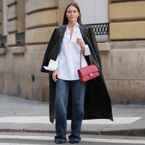 PARIS, FRANCE – DECEMBER 18: Diane Batoukina wears straight brown hair worn loose and parted at the center, clear eyeglasses, a white cotton button-up shirt, a long black leather coat, dark blue wide-leg denim jeans, black pointed-toe leather shoes, a red leather Chanel bag, during a street style fashion photo session, on December 18, 2025 in Paris, France.