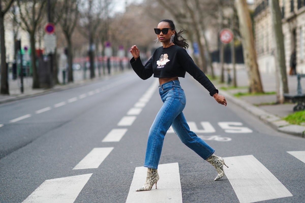 Emilie Joseph wears sunglasses, Isabel Marant bejeweled long earrings, a cropped black pullover with printed angels from Fiorucci, blue ribcage Levi's denim jeans, white high heels pointy ankle boots with snake print from Paris Texas, on February 16, 2021 in Paris, France.