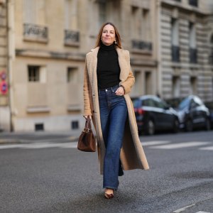 Alba Garavito Torre wears long brown hair parted at the center with natural skin and neutral lipstick, a black long-sleeve turtleneck pullover by Citizens of Humanity, dark-indigo high-rise flare jeans pants by Citizens of Humanity, a beige long shearling coat jacket by Possery, brown patent-leather shoes by Jonak, a brown suede New York midi bag by Demellier London, gold pearl earrings and gold rings by Sempiterno Paris, during a street style fashion photo session, on December 04, 2025 in Paris, France, full body shot.