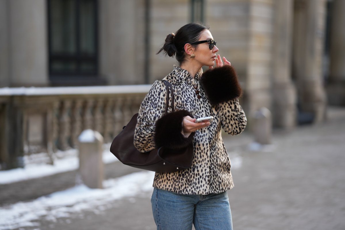 Milena Karl is seen wearing a cream and black leopard print "dolly" faux fur jacket with a classic collar, front leather button closure and dark brown fur trimmed cuffs from Carelli; light blue straight leg jeans with a mid rise fit from Massimo Dutti; the “Soft Margaux 15” handbag crafted in smooth dark brown leather with a softly structured silhouette, side buckle detailing, curved top handles and subtle logo detailing from The Row; holding an iPhone from Apple; rectangular black "Wayfarer" sunglasses with dark tinted lenses from Ray-Ban; sculptural gold colored earrings from Mango; her dark brown hair styled in a sleek low bun on February 14, 2026 in Hamburg, Germany.