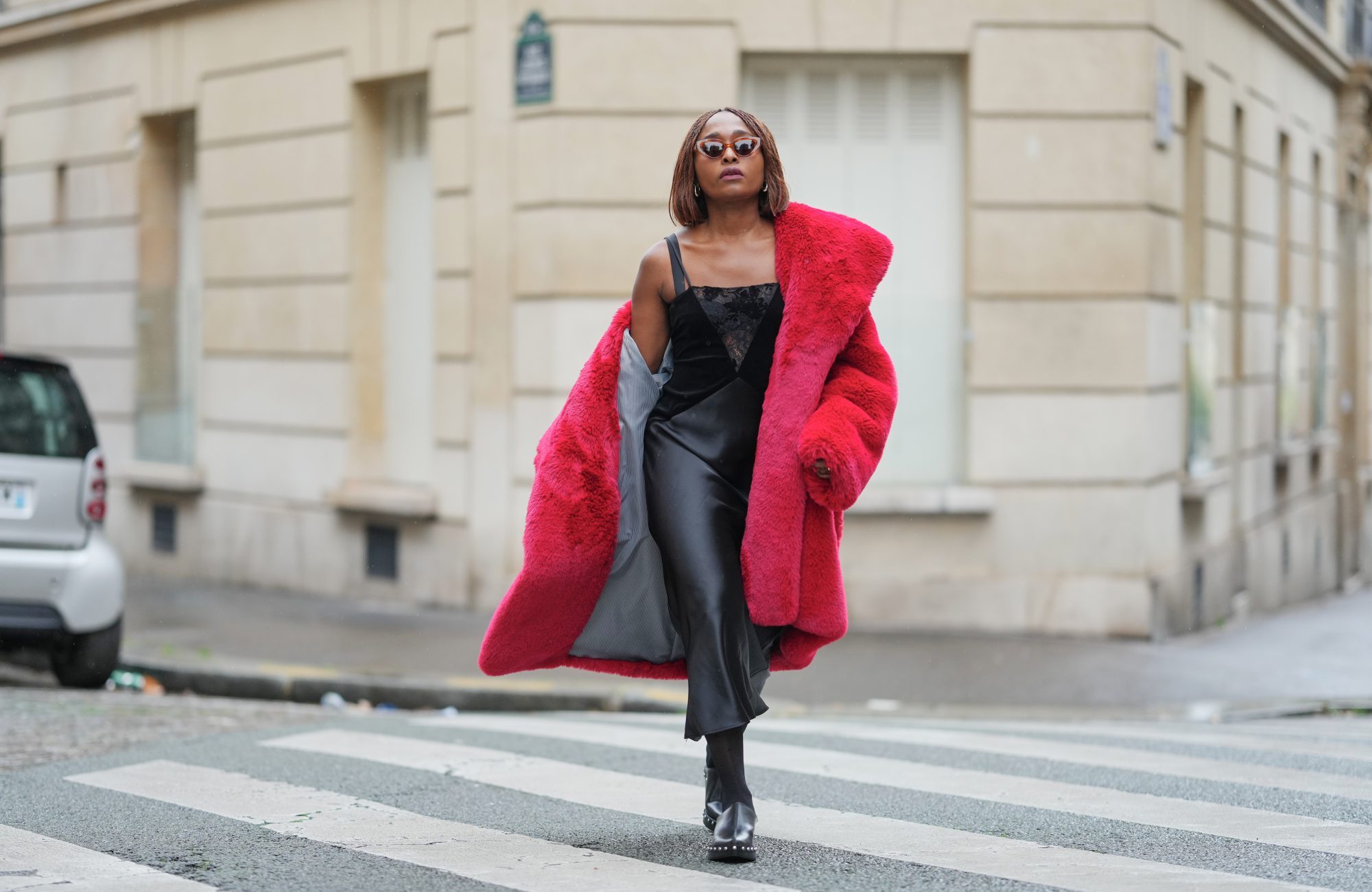 PARIS, FRANCE - DECEMBER 18: Carrole Sagba wears shoulder-length straight brown bob hair with a center part, makeup with an even complexion, defined brows and deep red lipstick, narrow brown-tinted sunglasses, a bright red faux-fur fluffy coat by Milkwhite worn open and draped off the shoulders, a black lace-trim camisole with thin straps, a black leather midi skirt with a straight cut and subtle sheen, sheer black tights, black leather clogs by Zara with a chunky sole, during a street style fashion photo session, on December 18, 2025 in Paris, France. (Photo by Edward Berthelot/Getty Images)