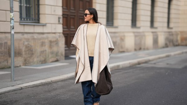 BERLIN, GERMANY - NOVEMBER 25: Laura Noltemeyer is seen wearing a cream oversized wool poncho with contrast whipstitch edging, a wide draped silhouette and open-front drape from Zara; layered underneath is a soft beige wool-blend knit sweater with a ribbed crew neckline and ribbed trims from Sézane; straight-leg dark-blue denim trousers with a classic fit from By Aylin Koenig; beige suede winter boots with a rounded toe, rope laces, tonal stitching and a thick rubber sole from Inuikii; a large textured dark-brown shoulder bag in a soft sculptural, slouchy shape and woven leather from Les Visionnaires x Michi from the Want; chunky sculptural gold earrings from Zara; black angular acetate sunglasses with a thick frame from Yves Saint Laurent; her dark brown hair is worn straight with a center part, tucked loosely behind the ears on November 25, 2025 in Berlin, Germany. (Photo by Moritz Scholz/Getty Images)
