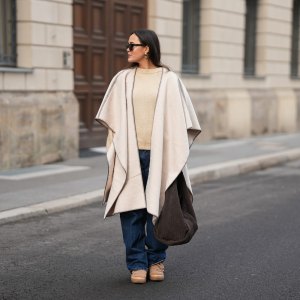 BERLIN, GERMANY - NOVEMBER 25: Laura Noltemeyer is seen wearing a cream oversized wool poncho with contrast whipstitch edging, a wide draped silhouette and open-front drape from Zara; layered underneath is a soft beige wool-blend knit sweater with a ribbed crew neckline and ribbed trims from Sézane; straight-leg dark-blue denim trousers with a classic fit from By Aylin Koenig; beige suede winter boots with a rounded toe, rope laces, tonal stitching and a thick rubber sole from Inuikii; a large textured dark-brown shoulder bag in a soft sculptural, slouchy shape and woven leather from Les Visionnaires x Michi from the Want; chunky sculptural gold earrings from Zara; black angular acetate sunglasses with a thick frame from Yves Saint Laurent; her dark brown hair is worn straight with a center part, tucked loosely behind the ears on November 25, 2025 in Berlin, Germany. (Photo by Moritz Scholz/Getty Images)