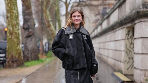 PARIS, FRANCE - JANUARY 25: Constance Jablonski wears long wavy blonde hair with a center part, metal hoop earrings, a handheld black leather clutch bag, a black oversized jacket with tonal PATOU logo-jacquard striping, textured leather collar and metal buckle details layered over a black top, a matching mini skirt, black glossy leather skinny boot, outside Patou, during Paris Fashion Week - Menswear Fall/Winter 2026-2027, on January 25, 2026 in Paris, France (Photo by Edward Berthelot/Getty Images)