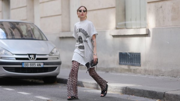 PARIS, FRANCE - JULY 17: Emy Venturini wears an oversized white graphic T-shirt dress by Balenciaga paired with flared leopard print trousers by Calzedonia. Black open-toe flat sandals by Prada complete the look. Accessories include a black leather clutch by Balenciaga, a silver Cartier wristwatch, gold sculptural earrings by Rabanne, and round sunglasses by Ray-Ban. Hair is blond and styled in a low ponytail with a center part, and lips are painted with lipstick in a bold red shade, during a street style fashion photo session, on July 17, 2025 in Paris, France. (Photo by Edward Berthelot/Getty Images)
