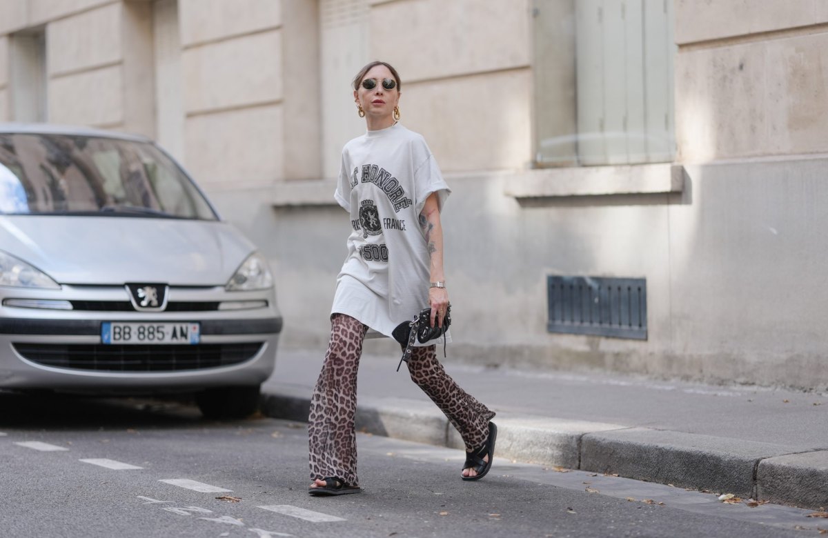 PARIS, FRANCE - JULY 17: Emy Venturini wears an oversized white graphic T-shirt dress by Balenciaga paired with flared leopard print trousers by Calzedonia. Black open-toe flat sandals by Prada complete the look. Accessories include a black leather clutch by Balenciaga, a silver Cartier wristwatch, gold sculptural earrings by Rabanne, and round sunglasses by Ray-Ban. Hair is blond and styled in a low ponytail with a center part, and lips are painted with lipstick in a bold red shade, during a street style fashion photo session, on July 17, 2025 in Paris, France. (Photo by Edward Berthelot/Getty Images)