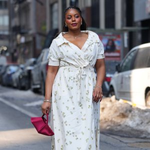NEW YORK, NEW YORK - FEBRUARY 14: A guest wears braided hair, earrings, necklace, gold rings, a pink satin clutch bag, a white V-neck wrap dress with a tie waist and a floral print, gold metallic open toe sandals shoes, outside Altuzarra, during New York Fashion Week, on February 14, 2026 in New York City (Photo by Edward Berthelot/Getty Images)
