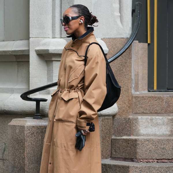 NEW YORK, NEW YORK - FEBRUARY 15: A guest wears black hair in a bun, black wraparound sunglasses, black wireless earbuds, a black woven backpack bag, a tan cotton high neck trench coat, black leather gloves, outside Diotima, during New York Fashion Week, on February 15, 2026 in New York City (Photo by Edward Berthelot/Getty Images)