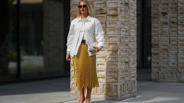 COPENHAGEN, DENMARK - AUGUST 06: A guest wears a white button-up shirt with a classic collar and long sleeves. The shirt is layered over a mustard yellow pleated skirt with intricate cut-out details near the hem. Sunglasses with dark lenses and a tortoiseshell frame are worn. A metallic clutch bag with a textured surface is held in one hand. The outfit is accessorized with a wide black belt featuring a prominent buckle. Footwear consists of silver strappy sandals with a low heel, outside Herskind, during Copenhagen Fashion Week, on August 06, 2025 in Copenhagen, Denmark (Photo by Edward Berthelot/Getty Images)