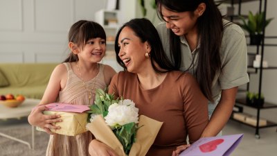 Heartwarming moment of two daughters giving their mother handmade cards, wrapped gifts, and a bouquet of flowers in a cozy living room. The family shares smiles and affection, celebrating Mother’s Day with love and appreciation. Perfect for concepts of motherhood, gratitude, family connection, and special occasions.