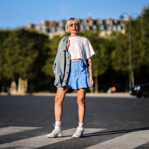 Emy venturini wears sunglasses from Prada, gold large earrings, a white cropped t-shirt from Ipsilon, a blue faded denim oversized long jacket, a blue with daisy flower print pattern flowing / high waist / knotted / short skirt from Zara, gold rings, white ribbed Calzedonia socks, white shiny leather Court Borough Mid 2 sneakers from Nike, on August 25, 2021 in Paris, France.