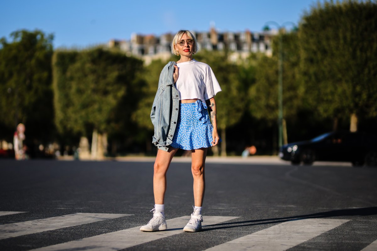 Emy venturini wears sunglasses from Prada, gold large earrings, a white cropped t-shirt from Ipsilon, a blue faded denim oversized long jacket, a blue with daisy flower print pattern flowing / high waist / knotted / short skirt from Zara, gold rings, white ribbed Calzedonia socks, white shiny leather Court Borough Mid 2 sneakers from Nike, on August 25, 2021 in Paris, France.