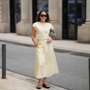 Laura Noltemeyer is seen wearing a yellow striped two-piece set with a sleeveless draped top and matching midi skirt by Sézane; black flip-flop sandals by Amazon; a black-and-white striped Porter clutch bag; black cat-eye sunglasses by Saint Laurent; nude-colored fingernails and toenails; gold jewelry by H&M and COS consisting of earrings, bangles and rings; natural makeup; her dark brown hair is worn in soft waves with a middle part on May 5, 2026 in Hamburg, Germany.