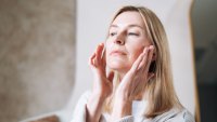 A focused adult woman practices facial self-massage in her well-lit bathroom. Using her fingertips, she gently smoothes her skin to improve circulation, reduce tension, and promote a healthy, natural glow. This authentic at-home wellness moment captures the popular beauty and self-care trend
