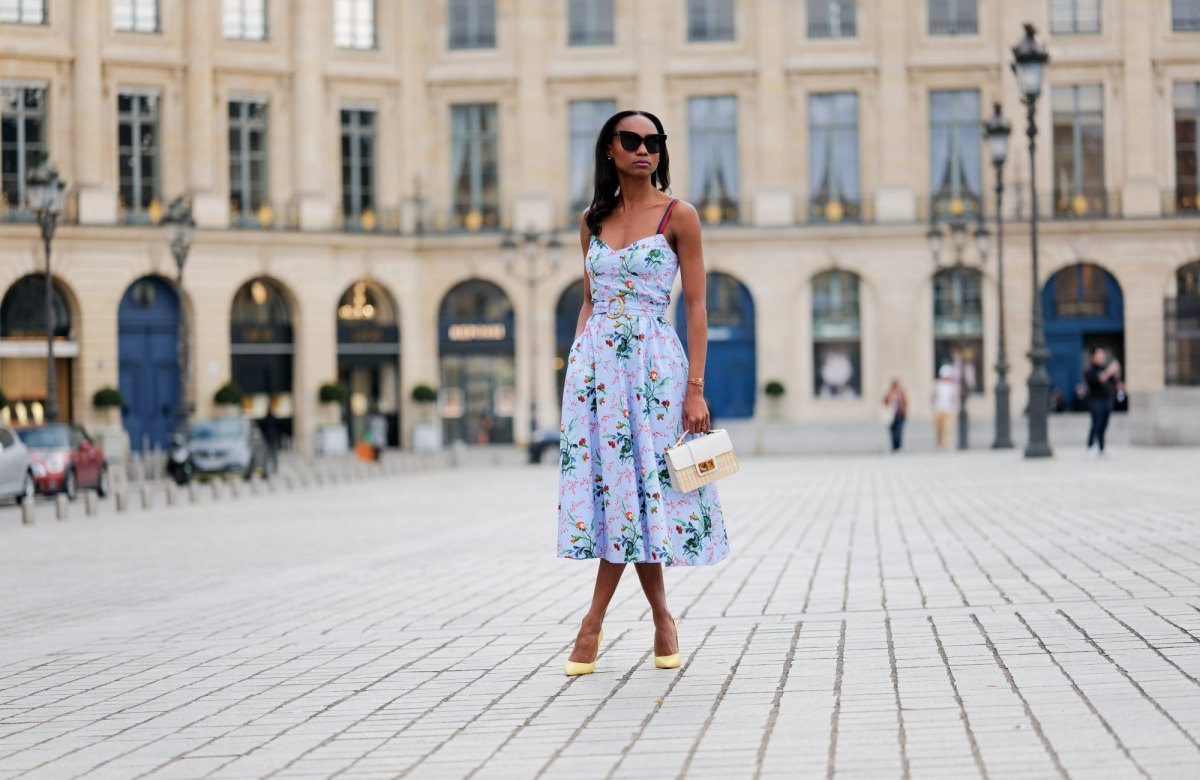 PARIS, FRANCE - APRIL 29: Emilie Joseph wears black large sunglasses, crystal earrings, a pale blue with red / green / navy blue / beige flower print pattern heart neck / tank-top / belted / midi dress, gold bracelets, a beige wickers and white shiny leather bi-material handbag, yellow shiny leather pointed pumps heels shoes , during a street style fashion photo session, on April 29, 2023 in Paris, France. (Photo by Edward Berthelot/Getty Images)