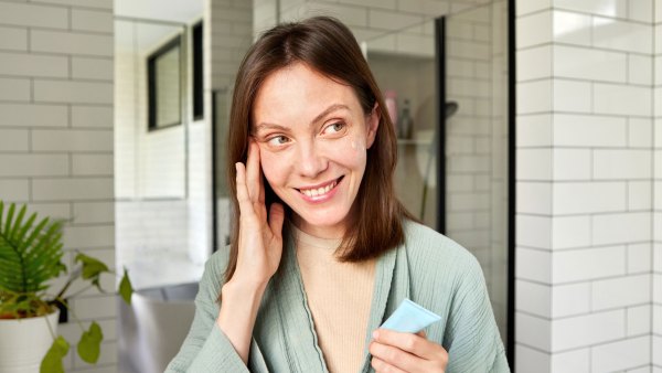Smiling young woman in a bathrobe applying moisturizing cream to her face after a refreshing shower in a modern bathroom, enjoying her daily self-care routine and promoting healthy, glowing skin