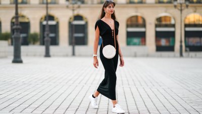 Amanda Derhy wears earrings, a black low-neck v-neck dress, a white circular round straw summer bag, a watch, white Adidas sneakers shoes, on August 09, 2020 in Paris, France.